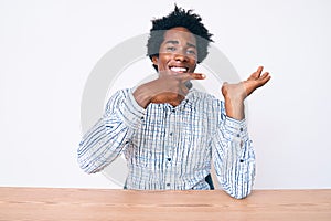 Handsome african american man with afro hair wearing casual clothes sitting on the table amazed and smiling to the camera while