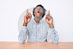 Handsome african american man with afro hair wearing casual clothes sitting on the table amazed and surprised looking up and