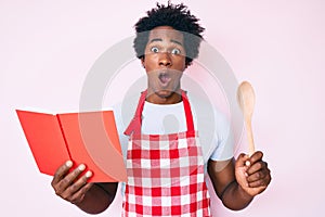 Handsome african american man with afro hair wearing professional baker apron reading cooking recipe book afraid and shocked with