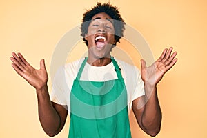 Handsome african american man with afro hair wearing waiter apron celebrating mad and crazy for success with arms raised and