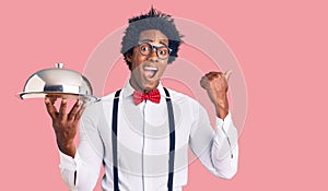 Handsome african american man with afro hair wearing waiter uniform holding silver tray pointing thumb up to the side smiling