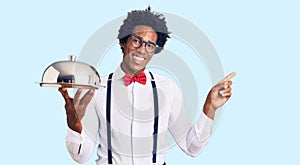 Handsome african american man with afro hair wearing waiter uniform holding silver tray smiling happy pointing with hand and