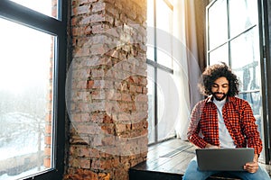 Handsome african american man with black beard works at the computer against the background of a large window