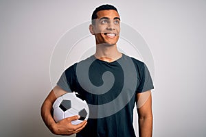 Handsome african american man playing footbal holding soccer ball over white background smiling looking to the side and staring