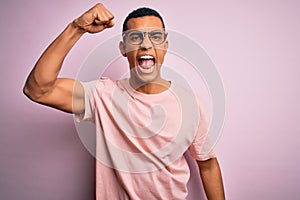 Handsome african american man wearing casual t-shirt and glasses over pink background angry and mad raising fist frustrated and