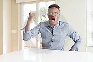 Handsome african american man on white table angry and mad raising fist frustrated and furious while shouting with anger