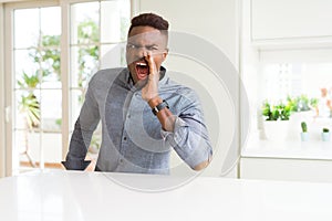 Handsome african american man on white table shouting and screaming loud to side with hand on mouth