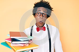 Handsome african american nerd man with afro hair holding books scared and amazed with open mouth for surprise, disbelief face