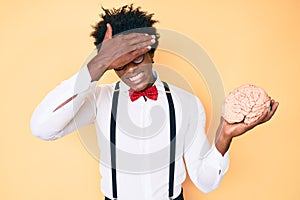 Handsome african american nerd man with afro hair holding brain stressed and frustrated with hand on head, surprised and angry