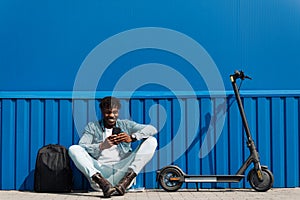 Handsome African American sitting on an electric scooter, with a phone in his hands, typing an SMS message, 5g internet