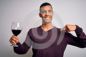 Handsome african american sommelier man tasting glass of red wine over white background with surprise face pointing finger to