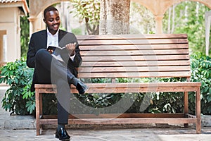 Handsome african businessman in a suit sits happily reading a book  on a chair in the park