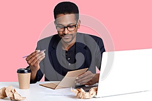 Handsome Afro American businessman in jacket and eyeglasses uses laptop. Black man holds pen and notebook in hands, wants to write