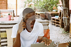 Handsome afro american man with glasses sitting at table in cafe on summer veranda, talking on telephone with closed