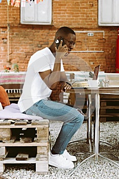 Handsome afro american man with glasses sitting at table in cafe on summer veranda, talking on telephone and working on