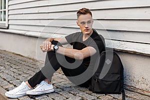 Handsome American man with a stylish hairstyle in a fashionable black T-shirt in stylish jeans in white sneakers with a backpack