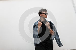 Handsome American young man with stylish hairstyle in denim fashionable clothes in trendy black sunglasses posing near a vintage