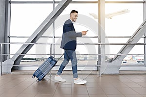 Handsome arab guy using smartphone while walking with suitcase at airport terminal