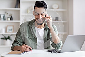 Handsome Arab Male Freelancer Talking On Cellphone And Taking Notes At Home