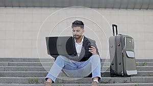 Handsome arabian man in stylish wear sitting on stairs and working on wireless laptop, while waiting for his flight