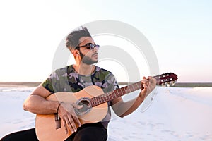 Handsome Arabian young man adjusts guitar, sitting on hill in mi