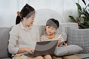 Handsome Asian boy, sitting on the sofa in the living room near his mother`s tablet, And his mother is working on a laptop