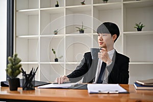 A handsome Asian male lawyer in a formal suit is sipping his morning coffee at his desk