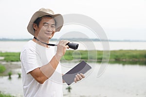 Handsome Asian man ecologist is surveying nature at the lake, holds binoculars and smart tablet. photo