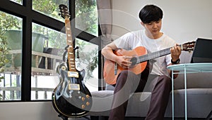 Handsome asian man playing guitar while sitting on couch in living room