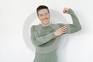 Handsome Asian man shows muscles posing after practising bodybuilding smiles happily wears casual green tshirt against white
