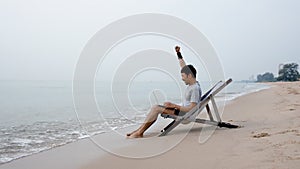 A handsome Asian man sits on a chair in a laptop computer by the sea as the waves crash against the shore, he raises his hand glad