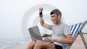 A handsome Asian man sits on a chair in a laptop computer by the sea as the waves crash against the shore, he raises his hand glad