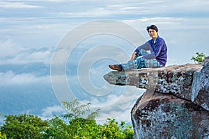 A handsome Asian man is sitting on a large rock that juts out from a steep cliff named Sut Phaen Din. Located at Pa Hin Ngam