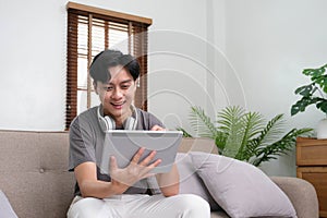 Handsome Asian man using digital tablet while headphones sitting on sofa at home