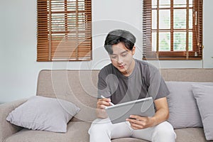 Handsome Asian man using digital tablet while headphones sitting on sofa at home