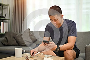 Handsome asian man using smart phone for making online payments, managing expenses finances in living room