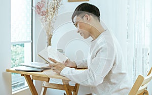 Handsome Asian man wearing glasses and casual clothes, reading book at desk or table beside window in bedroom at comfortable home