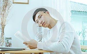 Handsome Asian man wearing glasses and casual clothes, reading book at desk or table beside window in bedroom at comfortable home