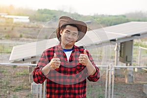 Handsome Asian man wears hat, red plaid shirt, stand beside solar panel at agriculture land. Thumbs up.