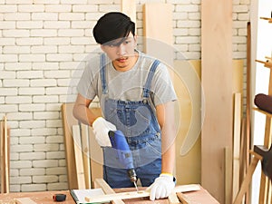 Handsome Asian repairman working in workshop with white brick background, drilling wood plank with electric drilling  machine