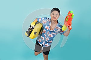 Handsome Asian tourist in summer clothes with water gun and luggage during Songkran festival on blue background