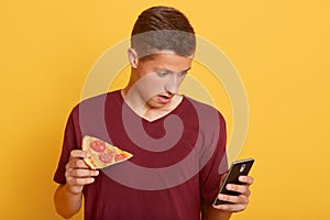 Handsome astonish guy holding slice of tasty pizza and phone, checking social network while having snack, sees braking news,