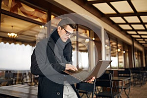 Handsome attractive businessman using a laptop on office terrace. Stylish man in glasses coat and scarf standing on