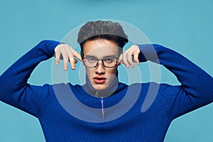 a handsome, attractive man stands on a light blue background in a blue zip-up sweater and holding his black-rimmed