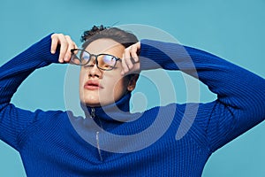 a handsome, attractive man stands on a light blue background in a blue zip-up sweater and holding his black-rimmed