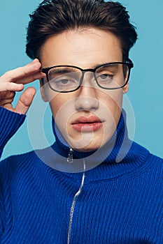 a handsome, attractive man stands on a light blue background in a blue zip-up sweater and holding his black-rimmed