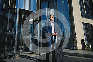 Handsome successful confident businessman in a business suit with black luggage against the background of a glass entrance to a