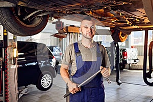 Handsome auto mechanic checking running gear of automobile on service station. Cheerful male worker fixing problem with car.