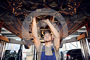 Handsome auto mechanic checking running gear of automobile on service station. Cheerful male worker fixing problem with car.