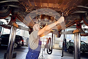 Handsome auto mechanic checking running gear of automobile on service station. Cheerful male worker fixing problem with car.